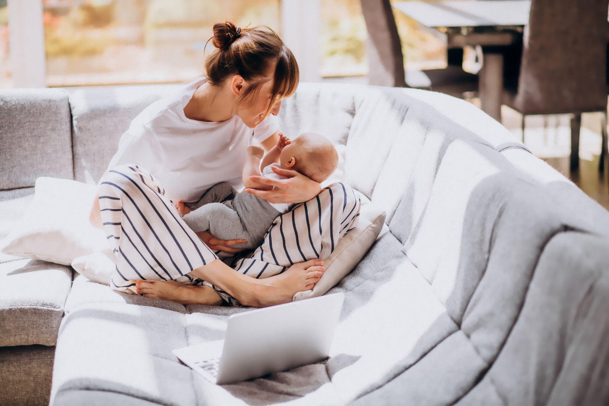 Junge Mutter, die mit Ihrem Baby auf einem Sofa sitzt. Daneben ist ein aufgeklapptes Laptop zu sehen.